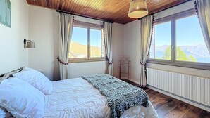 Een slaapkamer met een houten plafond, een bed met wit beddengoed, een gebreide plaid en twee ramen met gordijnen.