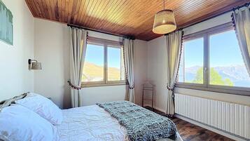 Een slaapkamer met een houten plafond, een bed met wit beddengoed, een gebreide plaid en twee ramen met gordijnen.