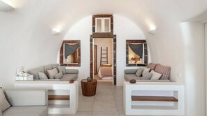 A loft-style room with a high ceiling, two sofas, a small wooden table, and a ladder leading to a lofted bed area. - Pura Vida Cave House (Santorini)