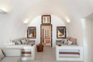 A loft-style room with a high ceiling, two sofas, a small wooden table, and a ladder leading to a lofted bed area. - Pura Vida Cave House (Santorini)