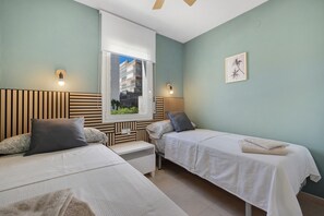 Ein Hotelzimmer mit zwei Betten, einem Fenster mit Blick auf Gebäude, einem Wandbild und einem Deckenventilator.