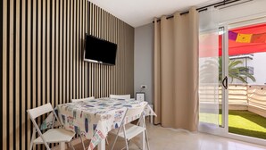 A room with a TV mounted on a striped wall, a table with a patterned tablecloth, two chairs, and a sliding glass door leading to an outdoor area with a red canopy. - Apartment 'Salou Beach' with Sea View, Wi-Fi, and Air Conditioning (Salou)