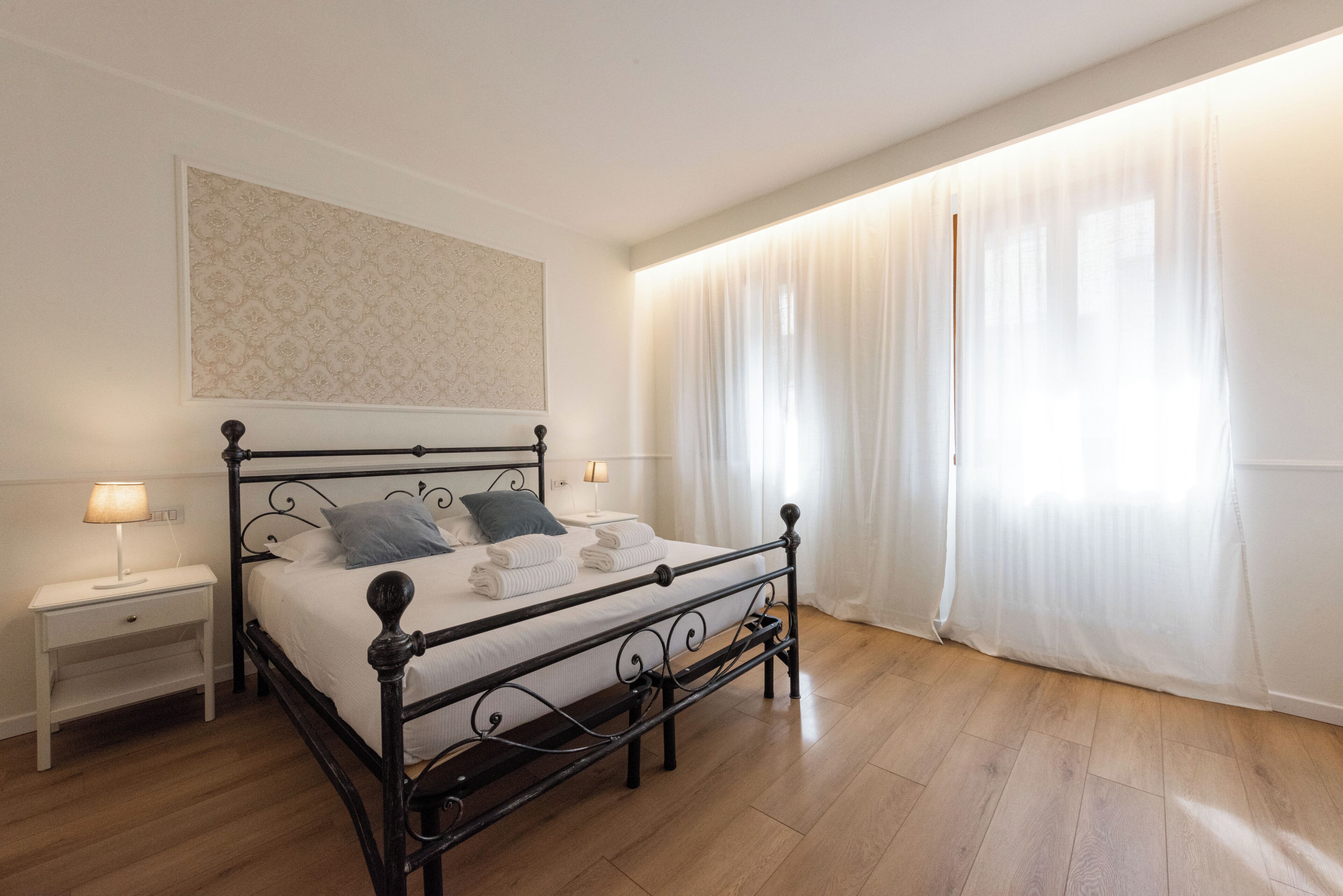 A neatly made bed with pillows, a bedside table with a lamp, and a large window with sheer curtains.