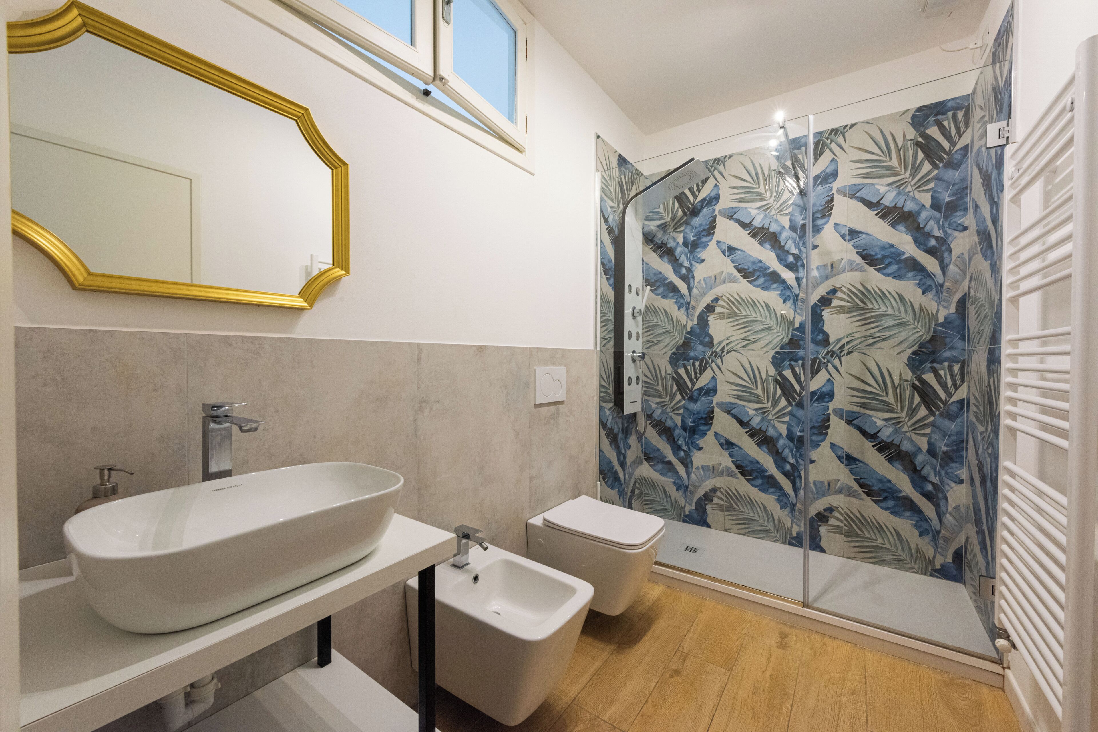 A bathroom with a large oval sink, a small bidet, a mirror with a gold frame, and a shower with a leaf-patterned glass door.