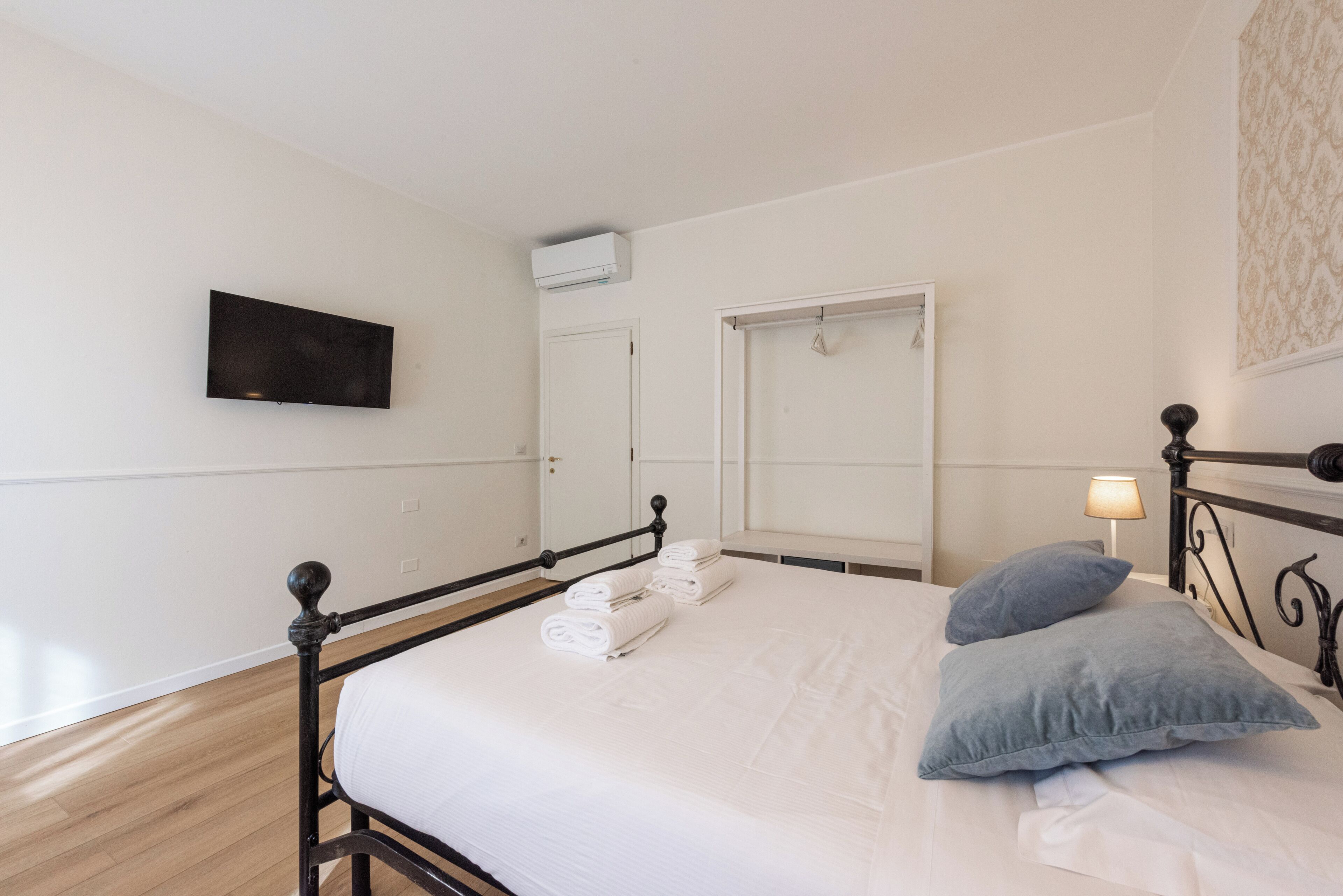 A neatly made bed with white linens and two pillows, a wall-mounted TV, a door with a mirror, and a small bedside lamp.
