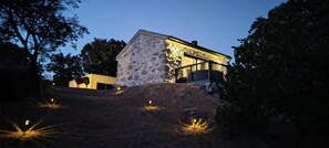 A stone house with a balcony, surrounded by trees and a sloped landscape.