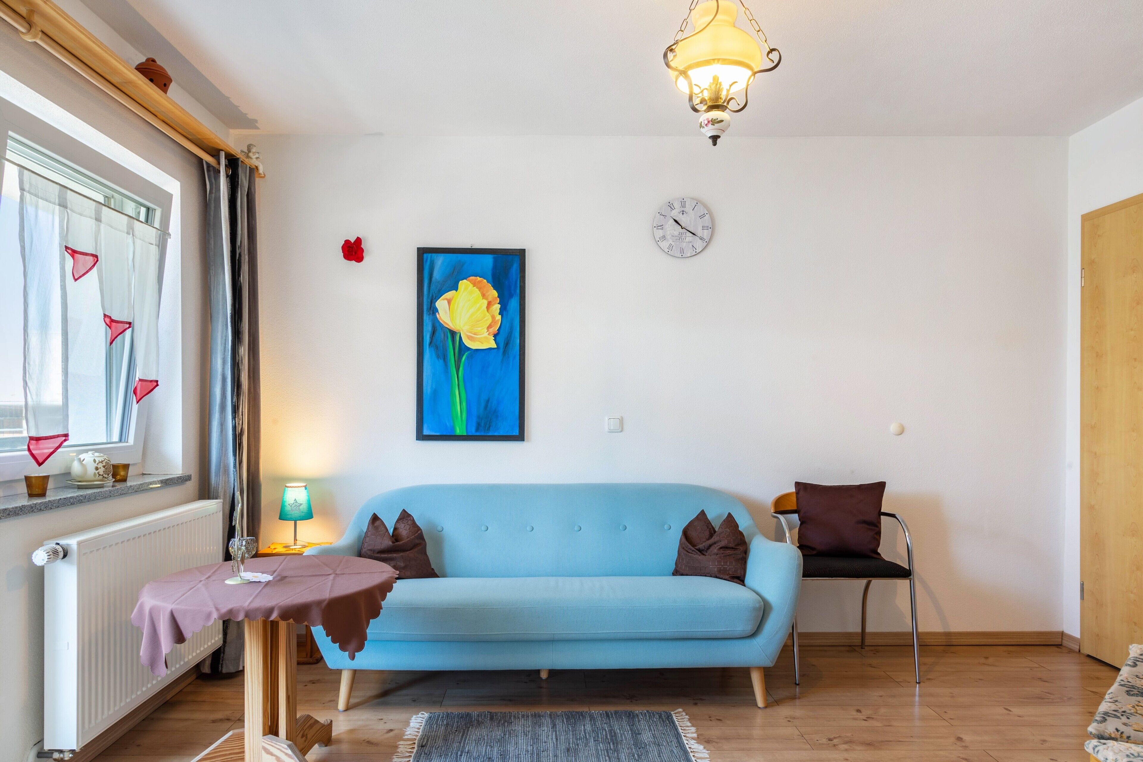 Una sala de estar con un sofá azul claro, una mesa con mantel morado, una lámpara, un cuadro de una flor, un reloj y una ventana con cortinas.