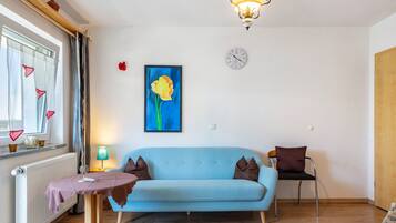 Una sala de estar con un sofá azul claro, una mesa con mantel morado, una lámpara, un cuadro de una flor, un reloj y una ventana con cortinas.