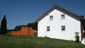 A white two-story house with a sloped roof, a wooden fence, and a grassy hill with dandelions.