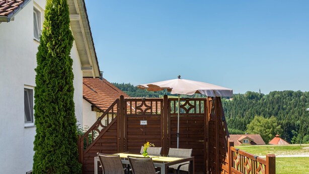 Eine Terrasse mit Tisch und Stühlen, ein Holzzaun und Blick auf ein bewaldetes Gebiet.