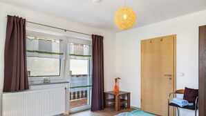 Ein Zimmer mit Bett, Stuhl, kleinem Tisch, Heizkörper, Tür und einem Fenster mit Vorhängen.