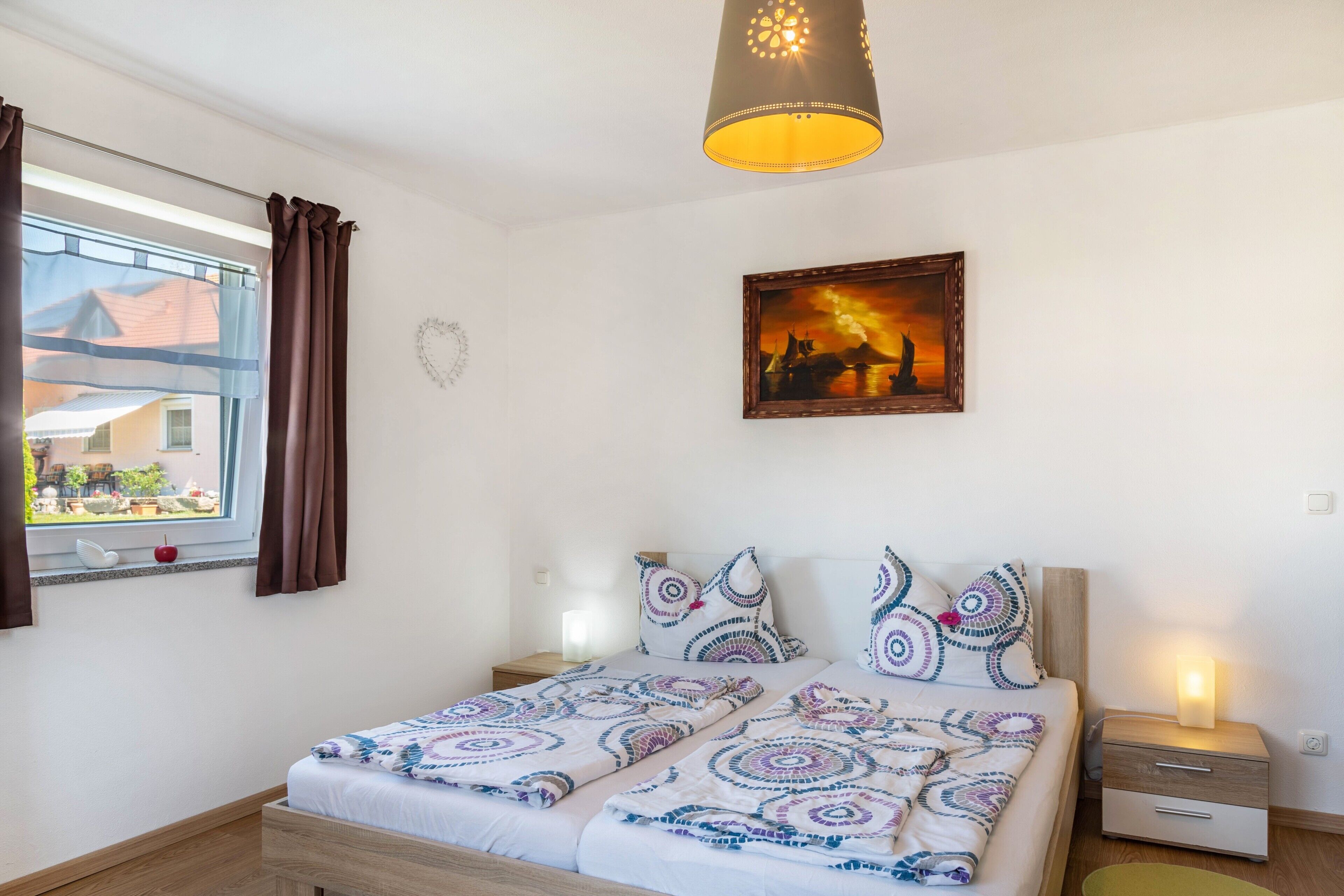 Un dormitorio con cama, mesita de noche, lámpara, ventana con cortinas y un cuadro en la pared.