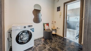 Ein Waschbereich mit einer Waschmaschine und einem Trockner, ein kleiner Hocker, eine Zierdeko und Blick durch eine Glastür in einen Küchenbereich.