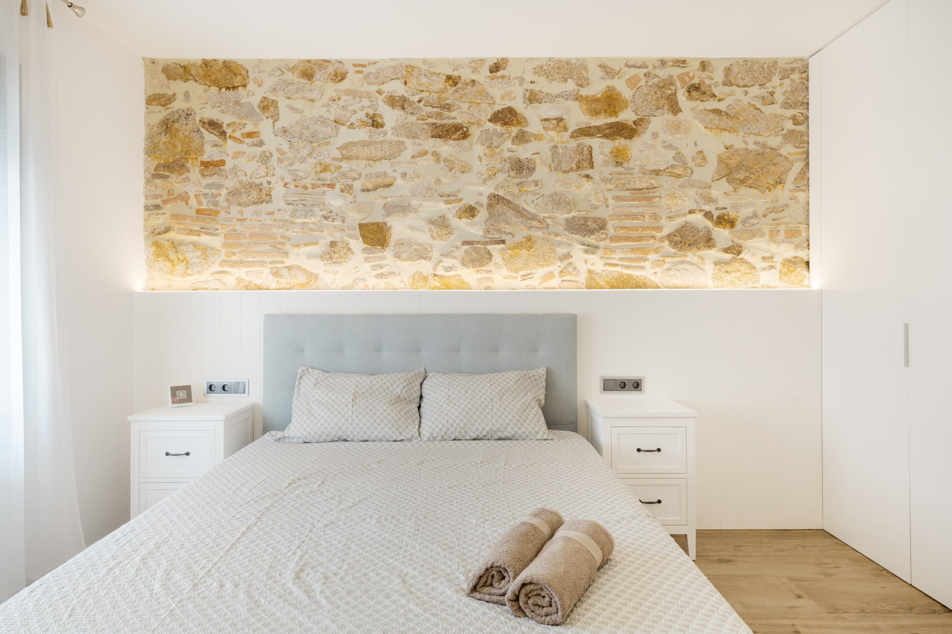Een slaapkamer met een stenen muurschildering, een gestoffeerd hoofdeinde, een wit sprei, twee nachtkastjes met lampen en een houten vloer.