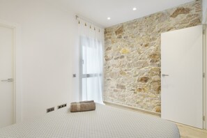 A bedroom with a stone wall, a bed with a patterned bedspread, a window with sheer curtains, and a white door.