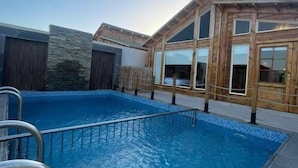 Una piscina con una escalera metálica, paredes exteriores de madera y grandes ventanales.