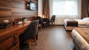 A hotel room with a wooden desk, two chairs, a bed, a TV mounted on the wall, and a window with curtains. - Zenith Hotel Frankfurt Airport  (Kelsterbach)