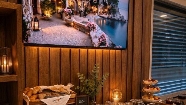 A breakfast buffet with various food items, a television displaying a scenic view, and a wooden wall panel. - Zenith Hotel Frankfurt Airport  (Kelsterbach)