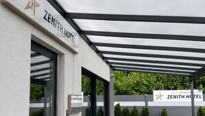 A modern hotel entrance with a covered outdoor area, a signboard, and a clear view of the surrounding greenery. - Zenith Hotel Frankfurt Airport  (Kelsterbach)