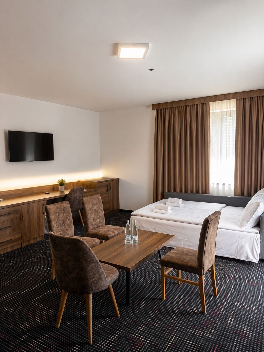 A hotel room with a bed, a desk with a flat-screen TV, two chairs, a table, and a patterned carpet.