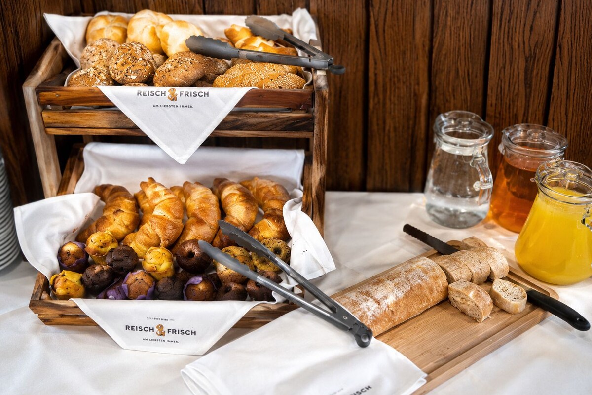 Un bufé de desayuno con panes y bollería variados, una tabla de cortar con rebanadas de pan y tarros de miel.