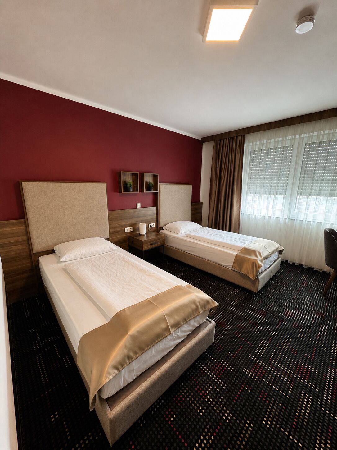A hotel room with two beds, a desk, a chair, a window with curtains, and a red wall.