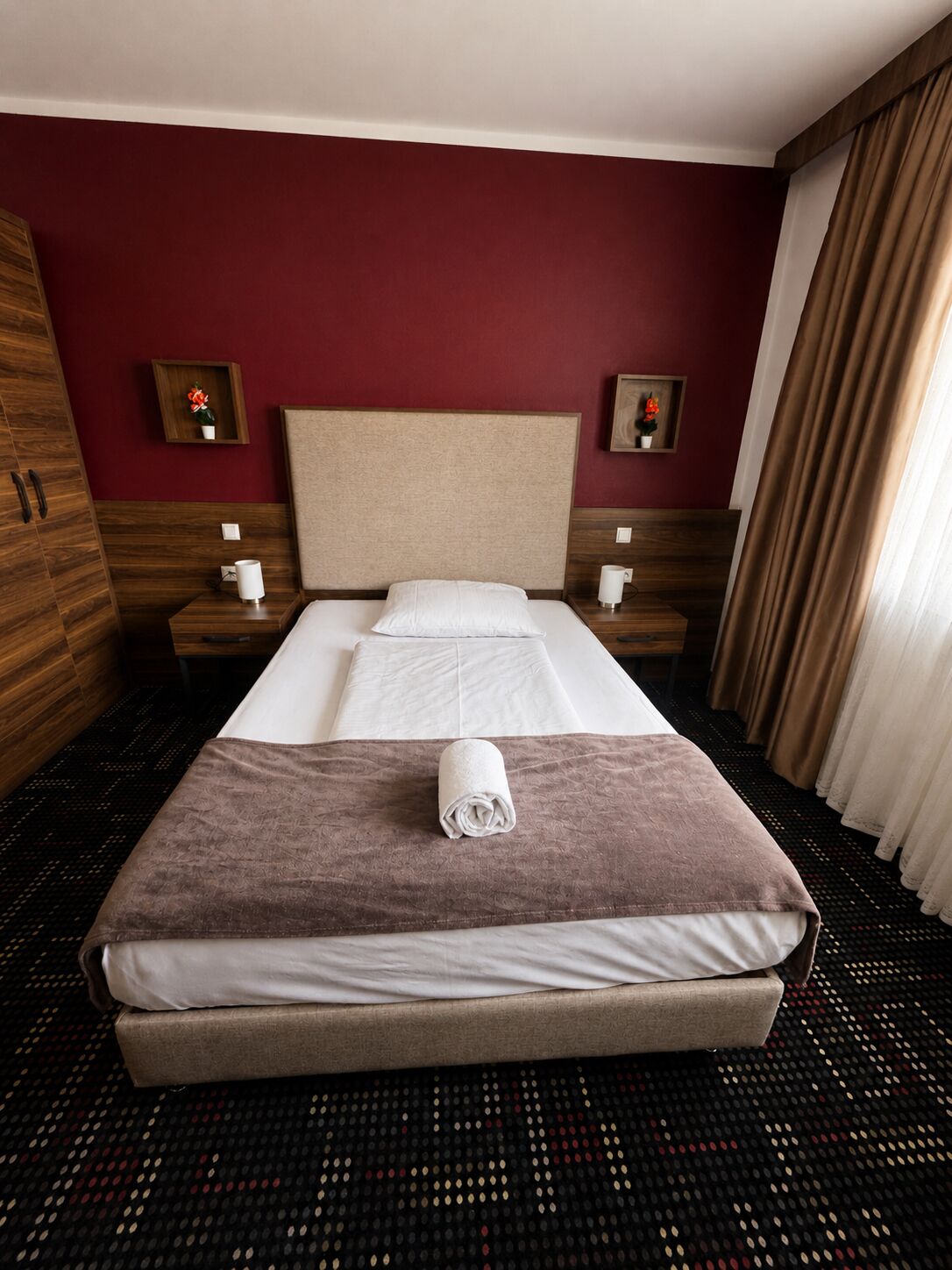 A hotel room with a bed, wooden wardrobe, two bedside tables, and a patterned carpet.
