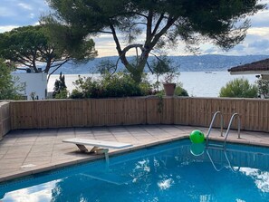 A swimming pool with a wooden deck, a diving board, and a view of a body of water and mountains. - Villa d’exception • Vue mer panoramique • Antibes (Antibes)