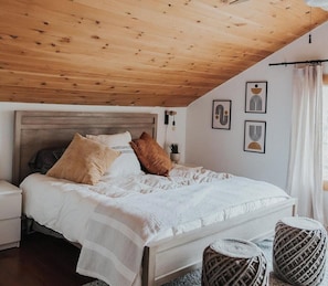 Un dormitorio con techo de madera, cama con ropa blanca, mesita de noche y decoración en la pared.