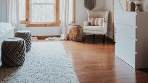 A cozy room with wooden ceiling beams, a ceiling fan, a large window with curtains, a white chair, a white dresser, and a woven rug. - Finger Lakes Renovated Farmhouse + Maple Farm! (Montour Falls)