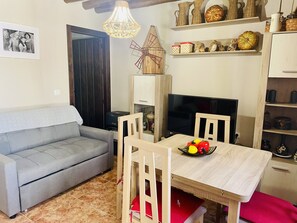 A living room with a grey sofa, wooden dining table, and chairs, a TV, a windmill decoration, and shelves with various items.