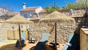 Une terrasse avec des chaises longues, un mur en pierre et une structure surmontée d’un toit de chaume.