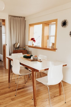 Un coin repas avec une table en bois et des chaises blanches, une fenêtre donnant sur un beau paysage et une petite étagère ornée d’objets décoratifs.