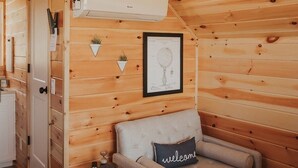 A cozy cabin interior with a wooden ceiling, a sofa with a 'welcome' pillow, a framed patent drawing, and a white air conditioning unit. - Tiny Cabin Stay in the Finger Lakes! (Science) (Burdett)