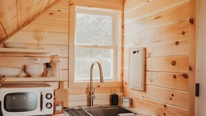 A compact kitchen with wooden walls, a window, a microwave, a sink, and cabinets. - Tiny Cabin Stay in the Finger Lakes! (Science) (Burdett)