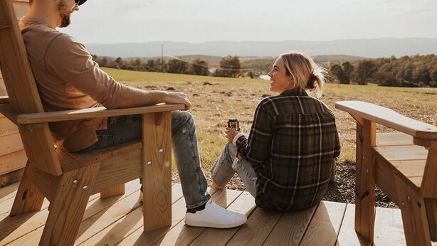 Two people sitting on wooden chairs on a wooden deck, facing each other with a view of a rural landscape in the background. - Tiny Cabin Stay in the Finger Lakes! (Science) (Burdett)
