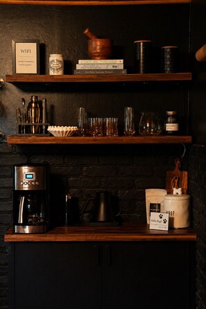 A kitchen area with a coffee machine, a kettle, and a coffee pot on the counter. There are shelves above with various items including a jar, a book, and a wooden bowl. - The Best Waterfalls Right Outside Your Door! (Montour Falls)