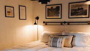 Una cama bien hecha con edredón blanco, una almohada estampada y un cabecero. Encima de la cama, en la pared, hay dos cuadros enmarcados.