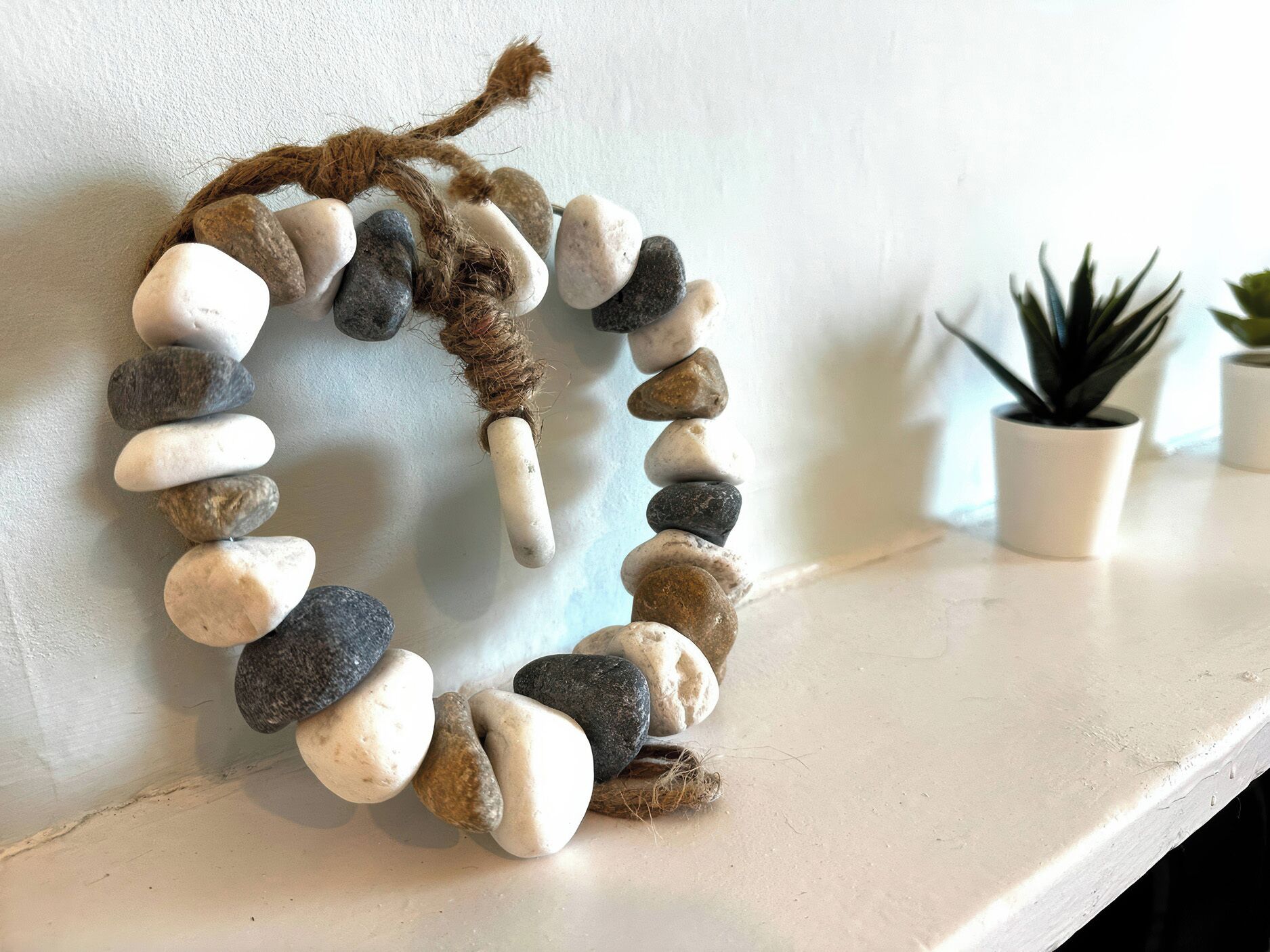 A wall-mounted arrangement of smooth, rounded stones of various sizes and colors, tied together with a natural fiber rope, set against a plain wall.