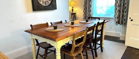 A dining room with a wooden table, six chairs, a wall clock, and a window with curtains.