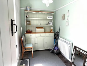 A small kitchen with a wooden chair, a shelf with various items, a radiator, and a vacuum cleaner. - Glanrafon (Llwyngwril)