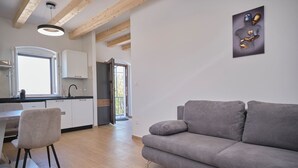 Ein modernes Wohnzimmer mit grauem Sofa, einem Essbereich mit weißem Tisch und Stühlen sowie einer Küche mit weißen Schränken und einem Fenster.