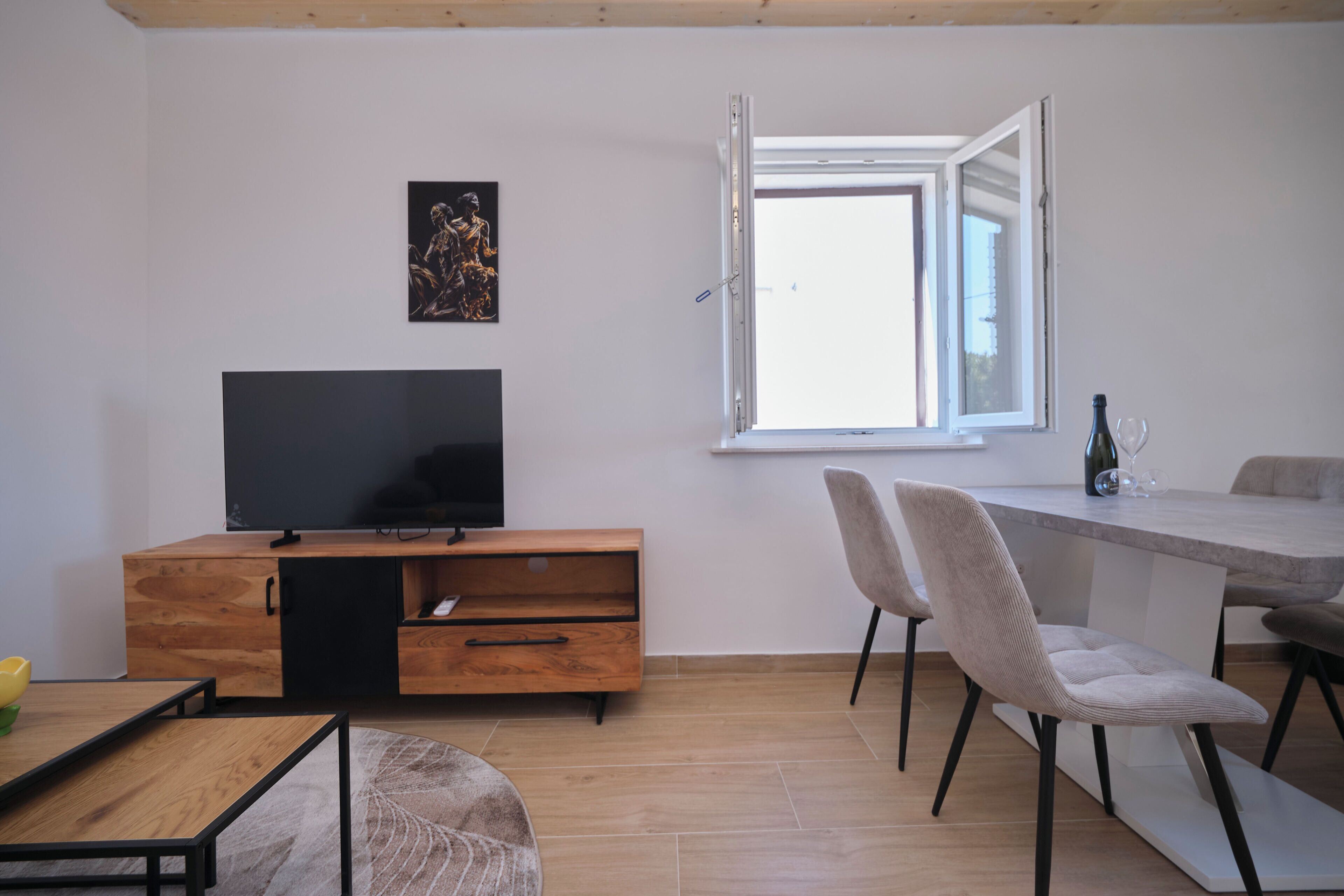 Ein modernes Wohnzimmer mit Flachbildfernseher, Holzmöbeln, einem Esstisch mit Stühlen und einem Fenster mit geöffnetem Klappladen.