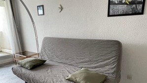 Un sofá gris con dos cojines, un marco de fotos y una lámpara de pie en una habitación con una pared lisa y una ventana.
