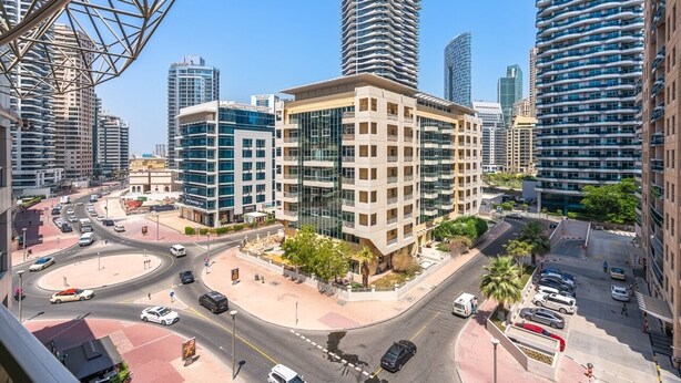 A modern urban street with high-rise buildings, a roundabout, and a parking area. - PVH - 2BR near Marina Promenade (Dubai)