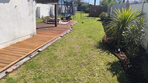 A backyard with a wooden deck, a patio table, chairs, a swing set, and a small garden area with plants and a palm tree. - House between Nantes and Pornic (Sainte-Pazanne)