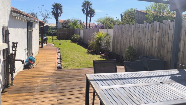 A wooden deck with a table and chairs, a garden with palm trees, and a wooden fence. - House between Nantes and Pornic (Sainte-Pazanne)
