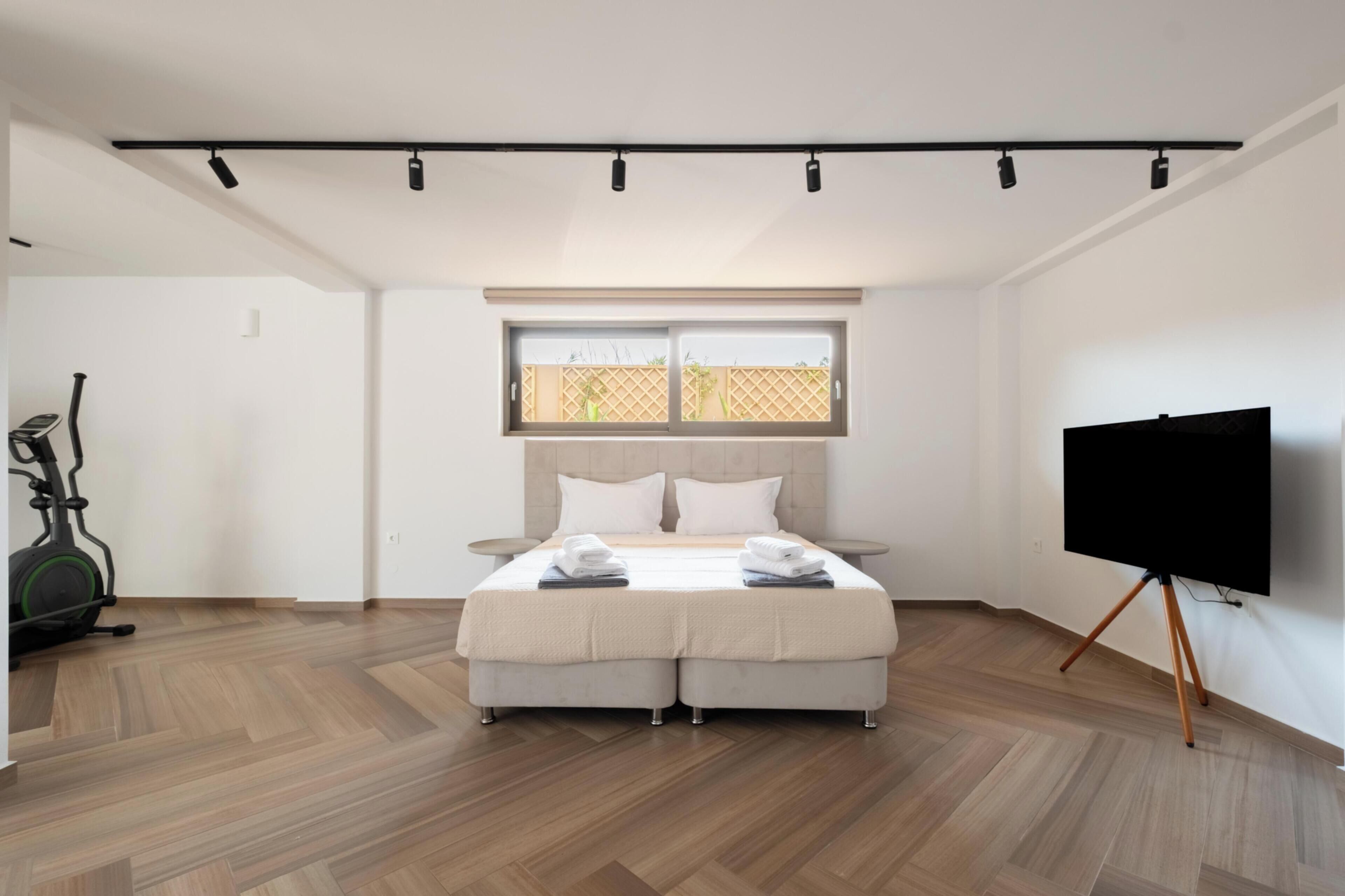 Quarto moderno com cama, televisão sobre suporte, janela e bicicleta ergométrica.