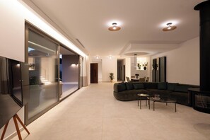 A modern living room with a sectional sofa, a glass wall, and recessed ceiling lights. - Villa Neratzia - Luxury Escape (Platanias)