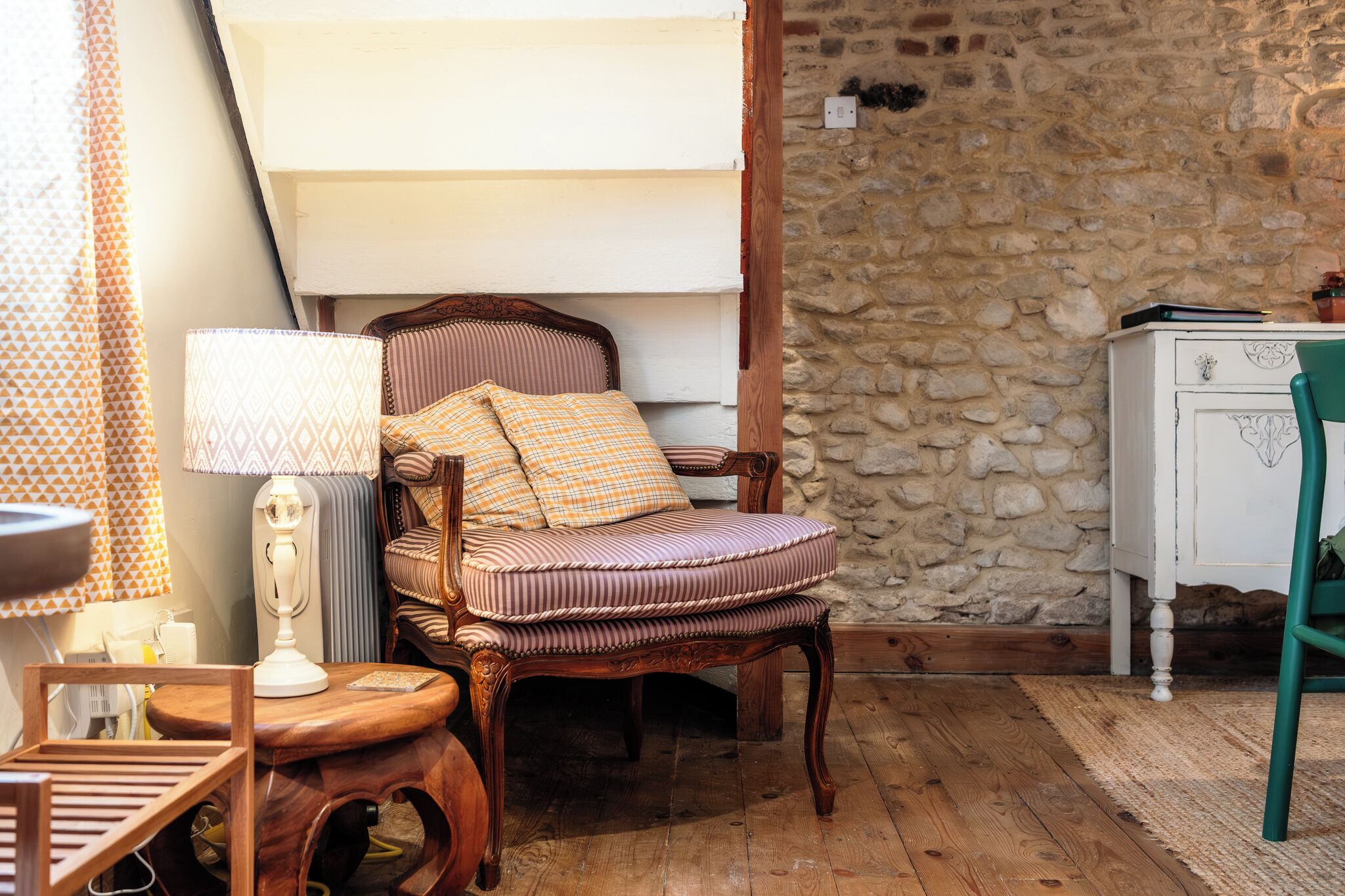 A rustic room with a wooden chair, a lamp, a small table, and a stone wall.